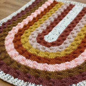 May include: A crocheted rainbow rug with a textured, bumpy surface. The rug is made in shades of brown, pink, yellow, and white.