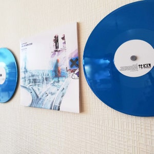 Peut inclure: Un disque vinyle bleu avec une étiquette blanche portant le texte "XL Recordings" et une petite illustration de deux personnages en bâton. Le disque est monté sur un mur blanc à côté d'une pochette d'album blanche avec le texte "OK Computer" et "Radiohead" imprimé dessus.