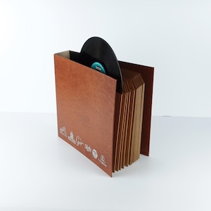 Puede incluir: Un estuche de almacenamiento de discos de vinilo de color marrón con aspecto de cuero, con un disco negro parcialmente insertado. El estuche tiene una serie de divisores y texto y gráficos plateados en la parte delantera. La etiqueta del disco es visible.