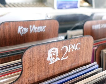 Puede incluir: Separadores de discos de madera con letras y gráficos blancos. Un separador dice "2PAC" con una imagen de perfil del artista. Otro dice "Kop Vicious". Los separadores están en una unidad de almacenamiento de discos de madera, con discos de vinilo visibles.