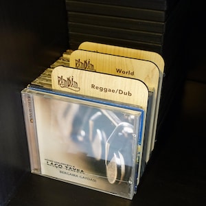 May include: A stack of CDs in clear plastic cases, neatly arranged with wooden dividers. The dividers are light wood, branded with "RamDam" and genre labels such as "Reggae/Dub" and "World." The CD cases display album artwork and text in a foreign language.
