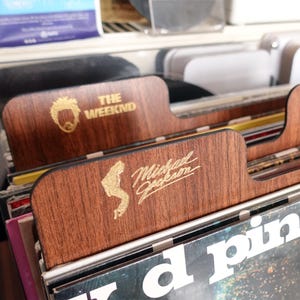 Op de afbeelding: Houten platenverdelers met gouden letters. Eén verdeler toont "THE WEEKND" en een gestileerd gezicht. Een andere toont "Michael Jackson" met een silhouet. De verdelers staan in een platenopslag.