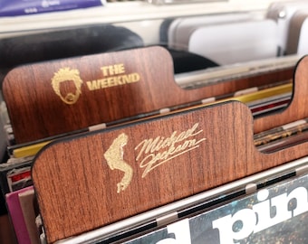 Puede incluir: Separadores de discos de vinilo de madera con letras doradas. Uno muestra "THE WEEKND" y una cara estilizada. Otro muestra "Michael Jackson" con una silueta. Los separadores están en un mueble para guardar discos.