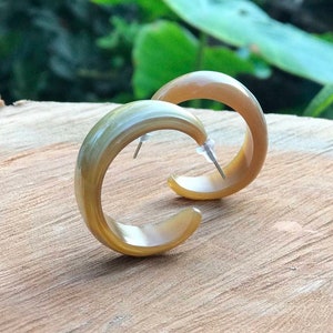 May include: A pair of hoop earrings made of natural horn. The earrings are a light brown color with a subtle green hue. They are shown on a wooden surface.