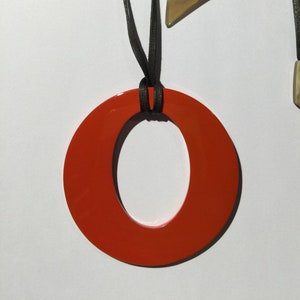 Puede incluir: Un collar moderno con un gran colgante circular naranja brillante suspendido de un cordón negro. El cordón tiene dos piezas geométricas en los extremos con un color marrón claro y blanco. El collar se muestra sobre un fondo blanco.