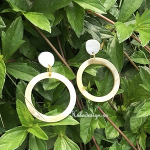 May include: A pair of white shell hoop earrings with gold accents. The earrings are photographed against a backdrop of green foliage.
