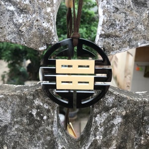 Op de afbeelding: Een zwarte en gouden ketting met een ronde hanger met een vierkant met vier lijnen erin. De hanger hangt aan een bruin koord.