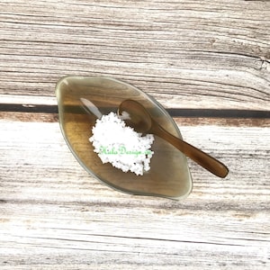 May include: A small, clear glass dish shaped like a leaf with a wooden spoon inside. The dish is filled with white salt crystals. The dish is sitting on a wooden surface.