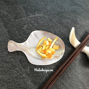 May include: A small, white, seashell-shaped dish with orange zest inside.  The dish is on a dark gray surface with a pair of dark brown chopsticks and a white seashell.