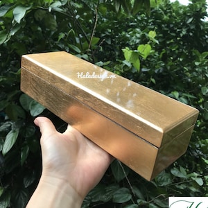 Może przedstawiać: Prostokątne pudełko w kolorze złotym z fakturowaną powierzchnią. Pudełko jest trzymane w dłoni.