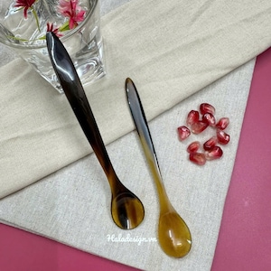 Puede incluir: Dos cucharas de cuerno marrón y negro sobre una tela de lino beige con semillas de granada rojas esparcidas a su alrededor.