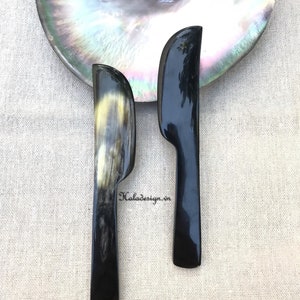 Puede incluir: Dos cuchillos de mantequilla de cuerno negro y marrón sobre una concha blanca e iridiscente. Los cuchillos miden aproximadamente 25 cm de largo.