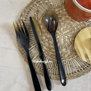 Könnte beinhalten: Ein Set aus drei schwarzen und braunen Hornbesteckteilen, bestehend aus einer Gabel, einem Messer und einem Löffel, auf einem geflochtenen Stroh-Untersetzer.