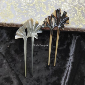 May include: Two decorative hair sticks with a ginkgo leaf design. The sticks are made of dark brown and light brown horn. The sticks are on a black velvet background.