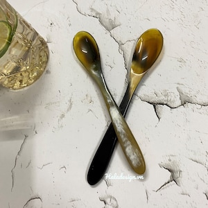 Puede incluir: Dos cucharas de cuerno, una negra y otra con un patrón de cuerno natural, se cruzan sobre una superficie blanca texturizada. Las cucharas tienen una forma de cuenco redondeada y un mango largo y delgado.