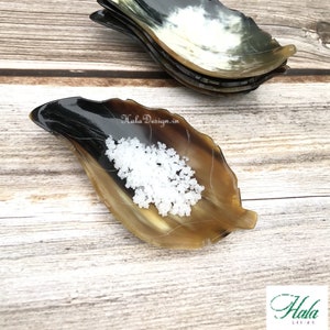 Op de afbeelding: Een set van drie kleine, blaadjesvormige schalen gemaakt van gepolijst hoorn. De schalen zijn op elkaar gestapeld en hebben een natuurlijke bruine en zwarte kleur. De bovenste schaal heeft een kleine hoeveelheid wit zout erin.