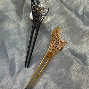 Può includere: Due forcine decorative per capelli. Una è nera, l'altra ambrata. Entrambe hanno un design traforato decorativo nella parte superiore e due lunghe punte. Gli accessori per capelli sono esposti su un tessuto azzurro.