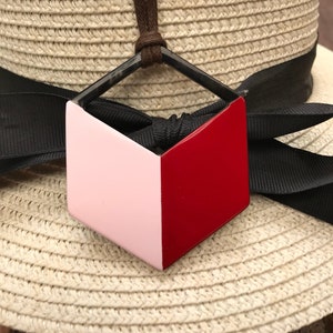 Puede incluir: Un collar con un colgante geométrico en un cordón negro. El colgante está hecho de dos colores: rosa y rojo. El colgante tiene la forma de un cubo con un lado cortado.