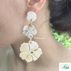 May include: A pair of white shell earrings with a flower design. The earrings are dangling and have a round disc at the top.
