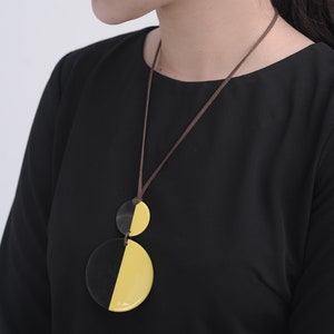 Puede incluir: Un collar negro y amarillo con un colgante hecho de dos semicírculos. El colgante está hecho de un material que parece carey. El collar lo lleva una persona con una camisa negra y pendientes amarillos y marrones.