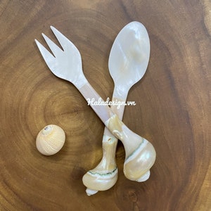 Könnte beinhalten: Ein Set aus zwei weißen Perlmuttlöffeln und einer Gabel mit braunen Griffen. Das Besteck ist in X-Form auf einer Holzoberfläche angeordnet. Die Griffe sind muschelförmig.