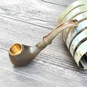 Puede incluir: Una pipa de cuerno marrón y beige con una banda de metal dorado y una cazoleta. La pipa está sobre una superficie de madera clara.