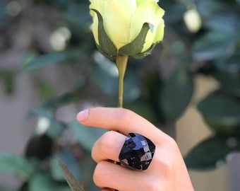 以下が含まれることがあります： 大きな、ファセット加工された黒い宝石の指輪。 指輪の幾何学的なデザインは光を反射し、きらめく効果を生み出します。 指輪は指に着用され、背景には淡い黄色のバラがあります。