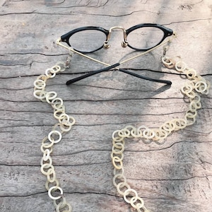 Könnte beinhalten: Schwarze Brille mit goldfarbenen Bügeln und einer cremefarbenen Kette aus verbundenen Kreisen. Die Kette liegt auf einer Holzoberfläche. Die Brille liegt auf der Holzoberfläche.