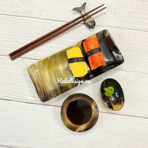 Op de afbeelding: Twee stukjes sushi op een bruin en zwart hoornen bord. Het ene stuk is geel en het andere is rood. Een klein schaaltje met groene wasabi en een klein schaaltje met donkerbruine sojasaus staan op een wit houten oppervlak. Een paar bruine eetstokjes liggen op een bruin en zwart hoornen visje.