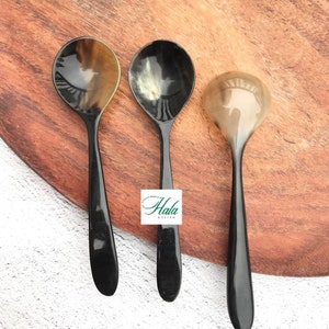 May include: Three black horn spoons with different patterns on the bowl. The spoons are on a wooden surface.
