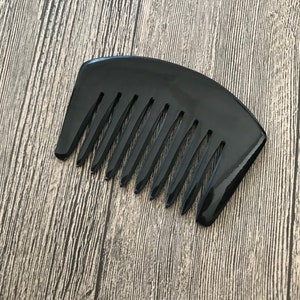 K&ouml;nnte beinhalten: Ein schwarzer Kamm mit breiten Z&auml;hnen und einem gebogenen Griff. Der Kamm ist aus einem gl&auml;nzenden, glatten Material.