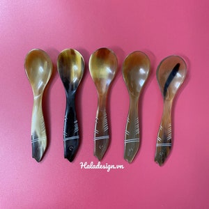 Puede incluir: Cinco cucharas pequeñas hechas de cuerno marrón con un diseño de pez tallado en el mango. Las cucharas están dispuestas en una fila sobre una superficie rosa.