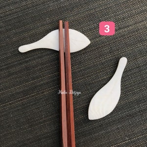 May include: A set of three wooden chopsticks and two white wooden spoons. The chopsticks are red and the spoons are shaped like fish. The text "Hola Design" is visible on the chopsticks.