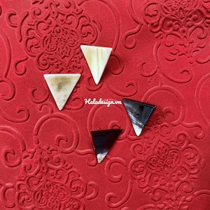 May include: Three triangle-shaped earrings made of horn. Two are light brown and one is black. The earrings are on a red background with a patterned design.