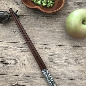 May include: A pair of brown wooden chopsticks with a decorative black and white mosaic design on the ends. The chopsticks are lying on a wooden surface.
