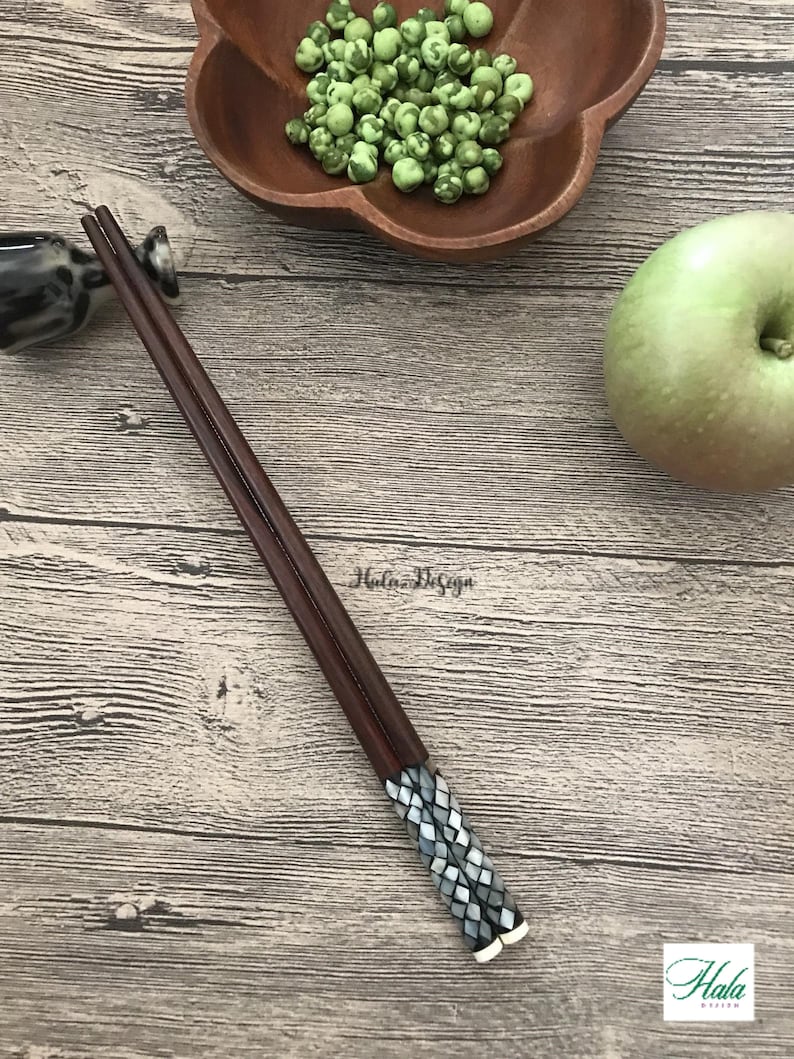 May include: A pair of brown wooden chopsticks with a decorative black and white mosaic design on the ends. The chopsticks are lying on a wooden surface.