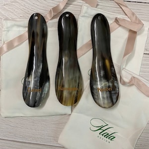 May include: Three brown horn shoe horns with gold lettering on white fabric bags. The bags have the text "Hala Design" printed on them.