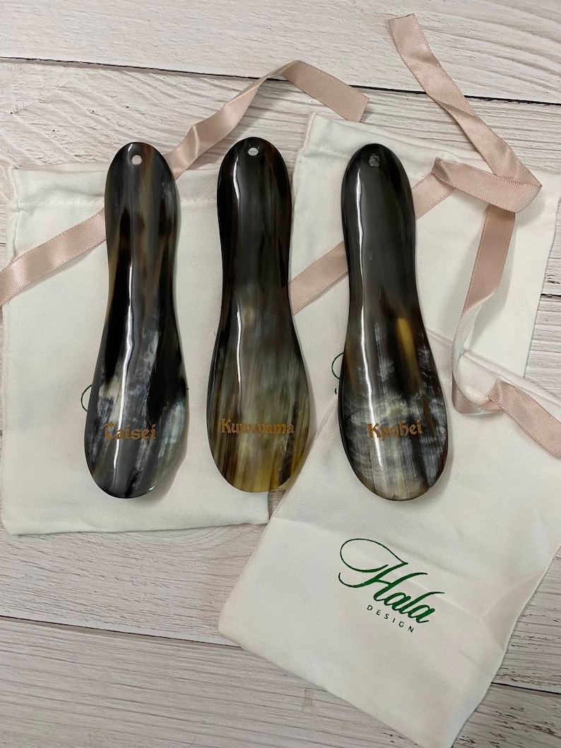 May include: Three brown horn shoe horns with gold lettering on white fabric bags. The bags have the text "Hala Design" printed on them.