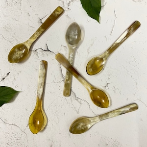 Puede incluir: Seis cucharas de cuerno marrón y blanco con cuencos redondeados. Las cucharas están dispuestas en forma de cruz sobre una superficie blanca texturizada.