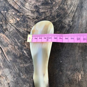 May include: A light brown horn shoe horn with a hole at the bottom. The horn is 5 inches long and is measured with a pink tape measure.