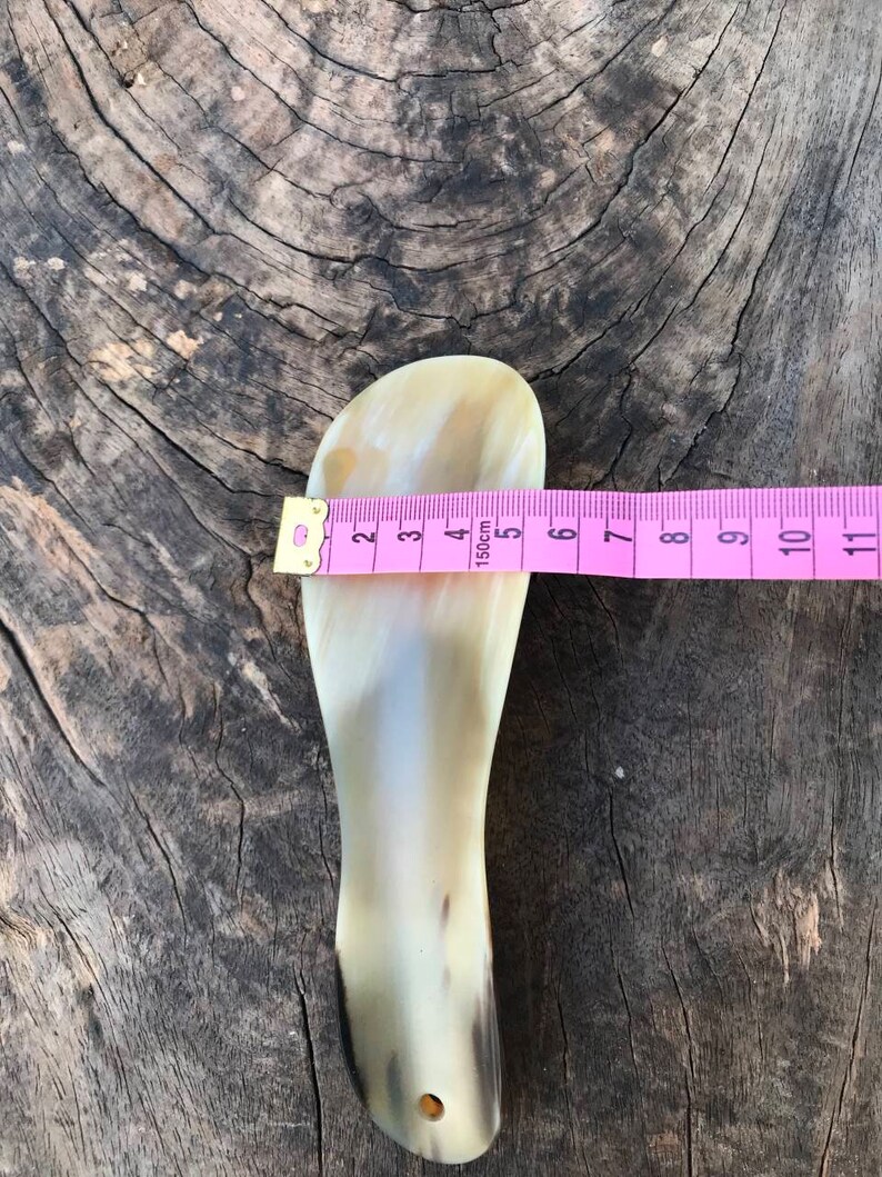 May include: A light brown horn shoe horn with a hole at the bottom. The horn is 5 inches long and is measured with a pink tape measure.