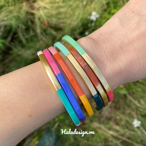 Könnte beinhalten: Ein Set bunter Armreifen mit goldenen Akzenten. Die Armreifen sind auf dem Handgelenk einer Person gestapelt und zeigen eine Vielzahl von Farben, darunter Türkis, Blau, Orange, Rosa und Grün.