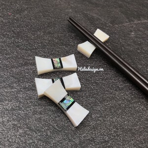 Könnte beinhalten: Drei weiße Perlmutt-Essstäbchen-Ablagen mit schwarzer und schillernder Einlage. Die Ablagen sind in Form einer Fliege geformt. Ein Paar dunkelbraune Essstäbchen liegen auf einer dunkelgrauen Oberfläche.