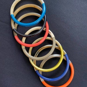 May include: A stack of seven horn bangles with different colored edges. The bangles are stacked in order of color, starting with blue, black, red, beige, gold, navy blue, and orange.