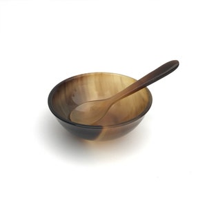 Pode incluir: Uma tigela de chifre marrom e bege com uma colher de chifre combinando. A tigela é redonda e tem um acabamento liso e polido.
