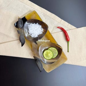 Könnte beinhalten: Ein Serviertablett mit zwei hornförmigen Schalen, eine gefüllt mit weißem Salz und die andere mit Limettenscheiben. Eine rote Chilischote liegt daneben. Das Tablett hat blattförmige Akzente und den Text "haladesign.vn".