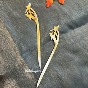 Könnte beinhalten: Zwei hellfarbene Haarstäbe mit dekorativen floralen Designs an der Spitze. Die Stäbe liegen auf einer dunkelgrauen, strukturierten Oberfläche. Ein farbenfrohes Blatt befindet sich im Hintergrund.