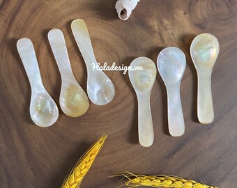 May include: Set of seven small, iridescent mother-of-pearl spoons on a wooden surface. The spoons are white with a pearlescent sheen and have a rounded bowl shape.