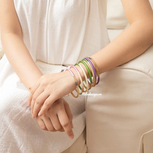 May include: A stack of colourful bangles on a person's wrist. The bangles are in shades of purple, green, gold, and white.