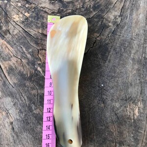 May include: A light brown horn shoe horn with a hole near the bottom. The horn is about 6 inches long and is measured against a pink tape measure.